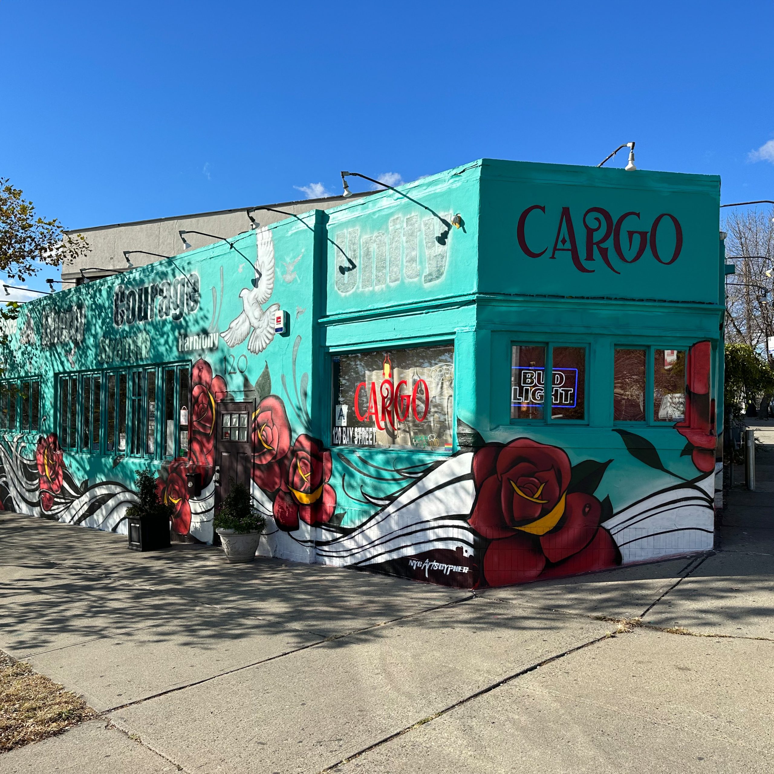Wide-angle shot of Cargo Cafe's exterior showcasing the NYC Arts Cypher's Fall 2023 mural, featuring large red roses and a dove on a teal wall, under a clear sky.